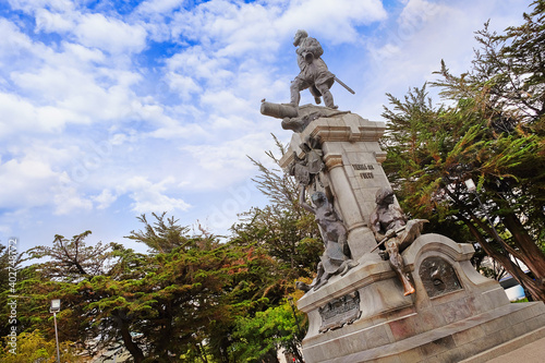 Monument of Ferdinand Magellan in the Plaza de Armas, Punta Arenas, Chile, surrounded by green vegetation, against a blue sky covered by white clouds.