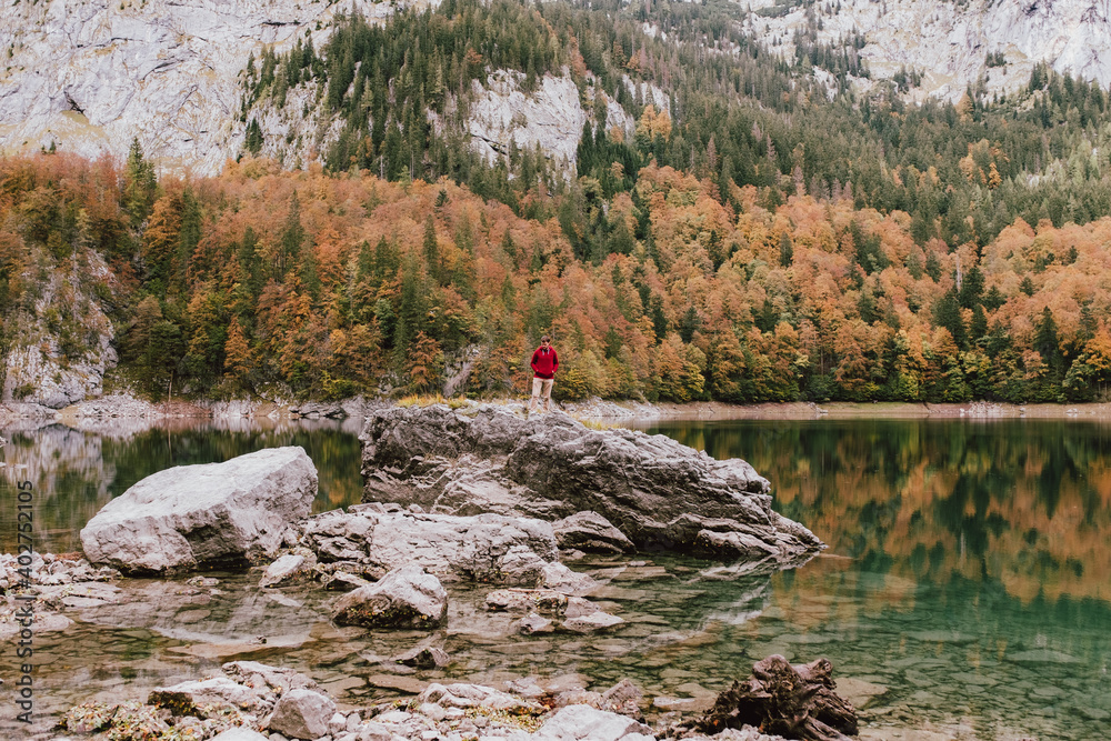 Man in a red sweater standing on a rock in the middle of a lake surrounded by autumn colors trees and mountain in the background