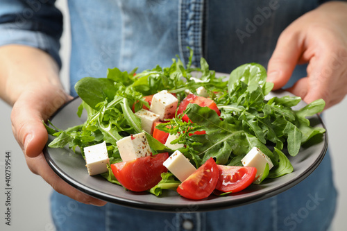 Woman holding plate of delicious salad with feta cheese, arugula and tomatoes on light background, closeup