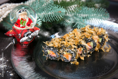 Japanese sushi roll on Christmas decorated plate. Raw salmon, tuna and cucumber wrapped in black rice with tuna chips Bonita on top. New year tree brunch and small Santa Clause statuette on background