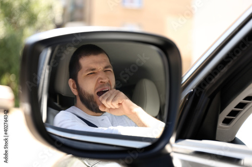 Canvas Print Tired man yawning in his modern car, view through car side mirror