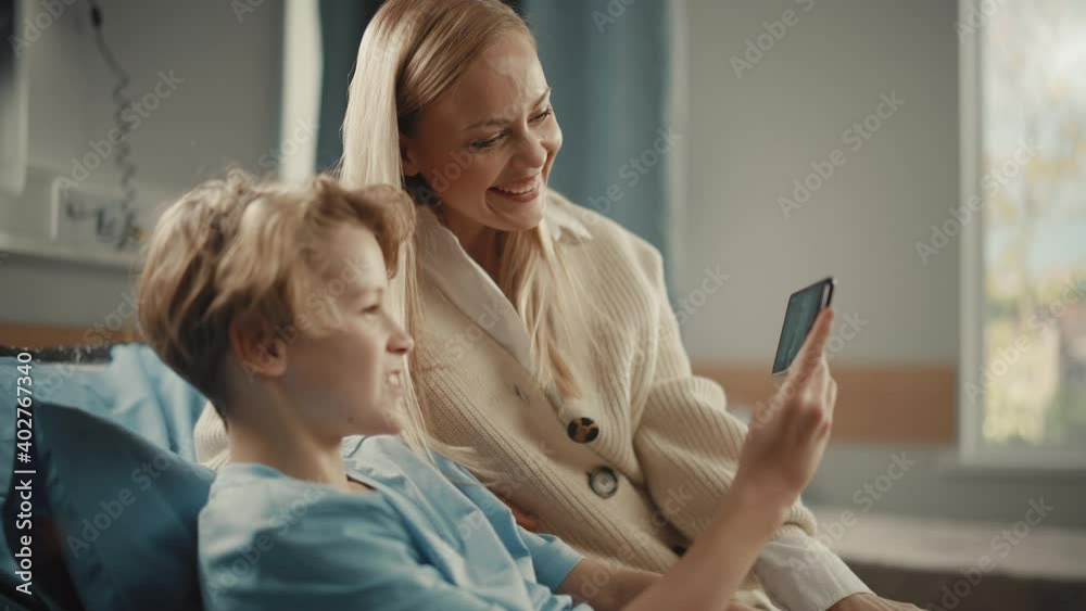 Hospital Ward: Handsome Young Boy Resting in Bed with His Beautiful Mother Visiting Uses Smartphone for Video Call Conference Talk with Family and Friends. Happy Patient Recovering after Sickness Hospital Ward: Handsome Young Boy Resting in Bed with His Beautiful Mother Visiting Uses Smartphone for Video Call Conference Talk with Family and Friends. Happy Patient Recovering after Sickness