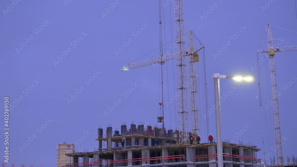 Engineers and builders in helmets are working on a monolithic construction site. A construction crane carries a load during a snowfall in the evening. Engineers and builders in helmets are working on a monolithic construction site. A construction crane carries a load during a snowfall in the evening.