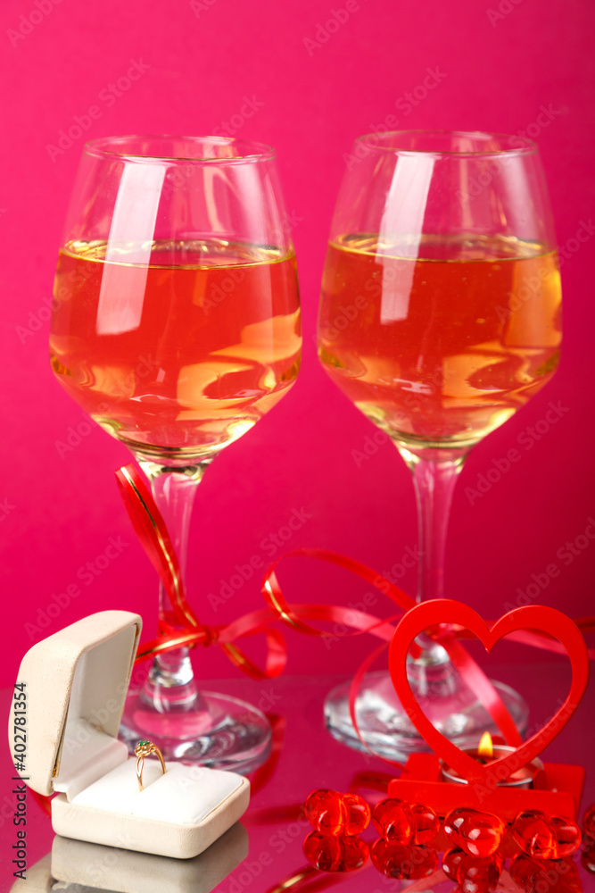 Two glasses of champagne tied with red ribbons on a pink background next to a ring in a box and a candle in a candlestick. Two glasses of champagne tied with red ribbons on a pink background next to a ring in a box and a candle in a candlestick.