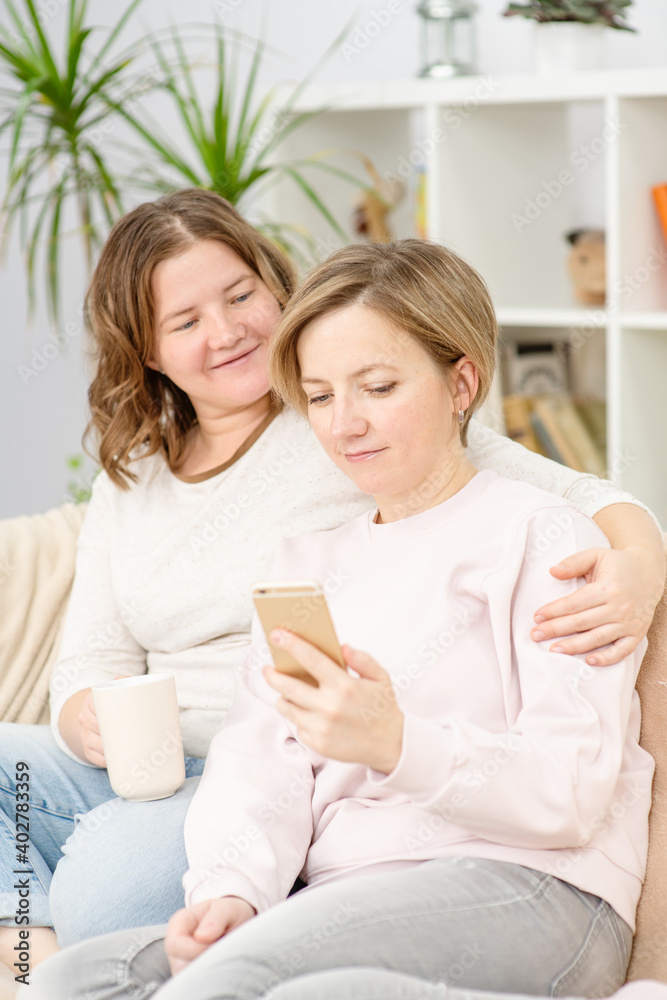 Beautiful young women LGBT lesbian happy couple sitting on sofa buying online using phone in living room at home. LGBT lesbian couple together indoors concept. Beautiful young women LGBT lesbian happy couple sitting on sofa buying online using phone in living room at home. LGBT lesbian couple together indoors concept.