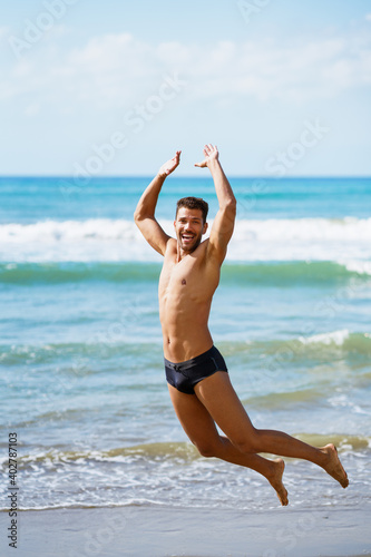 Young man with beautiful body in swimwear jumping on a tropical beach.