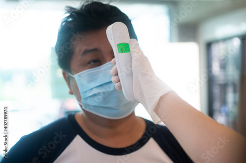 Asian waitress woman wearing face masks and holding an infrared forehead thermometer to check body temperature for virus symptoms of customers before entering the restaurant ( coffee shop ).