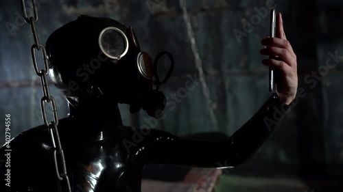 A passionate woman dressed in a black latex suit for a BDSM role-playing game, a gas mask on her face, she wants a selfie on her smartphone, is on a bed with chains.