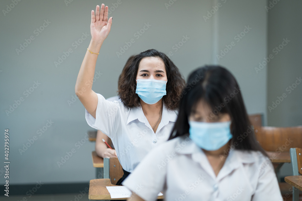 College students wear facemask study in classroom and raise hand to ask teacher during coronavirus pandemic. Selective focus portrait of university students study in the classroom. Students in uniform