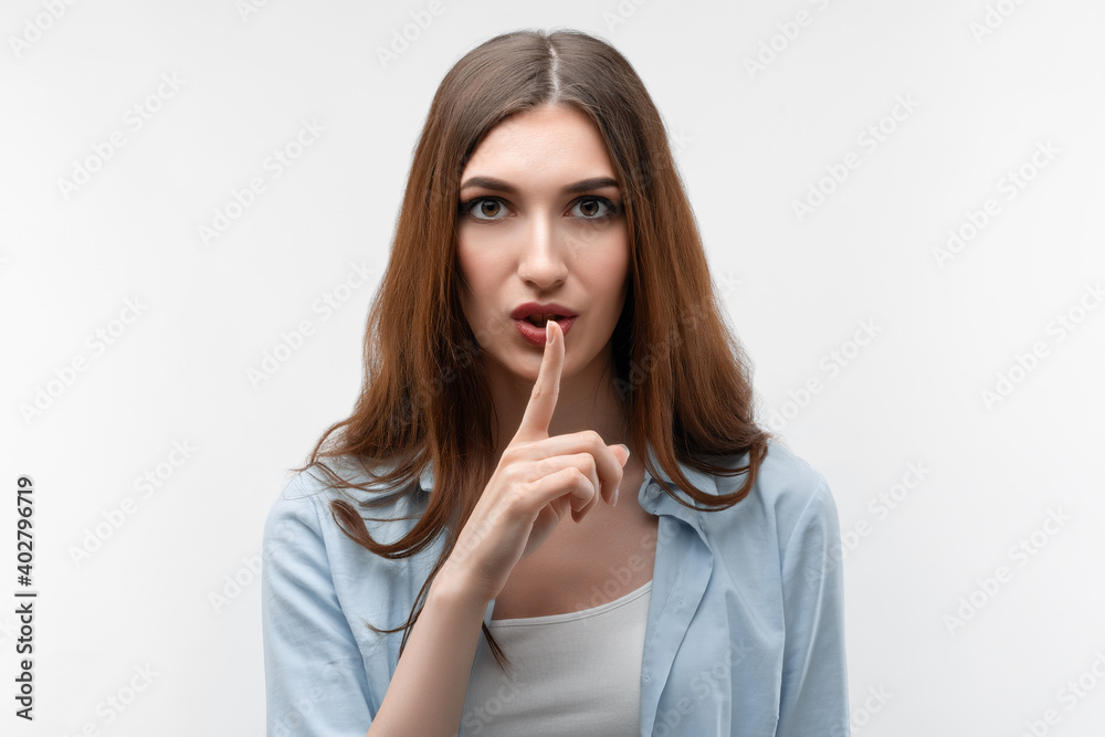 Pretty girl with long chestnut hair, dressed in casual clothes, keeps finger on lips, making hush gesture on white background. Shh, silence concept Pretty girl with long chestnut hair, dressed in casual clothes, keeps finger on lips, making hush gesture on white background. Shh, silence concept