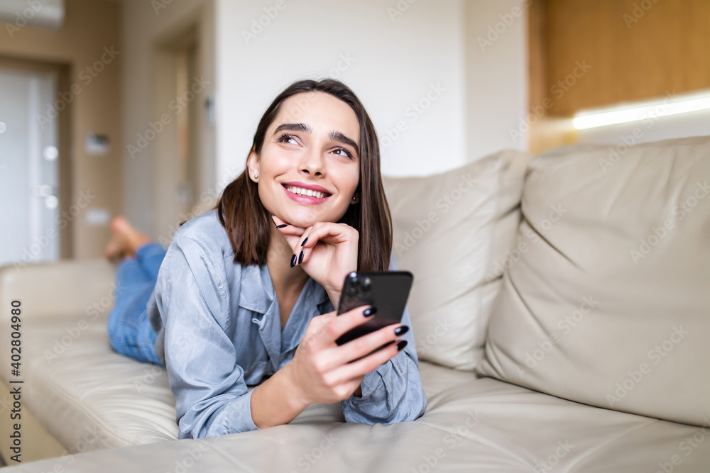 Portrait of dreamy thoughtful woman lying on stomach on couch pillow holding smart phone in hands looking away Portrait of dreamy thoughtful woman lying on stomach on couch pillow holding smart phone in hands looking away