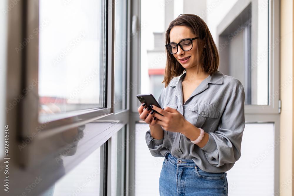 Side view portrait of a casual woman using a smart phone near a window of the living room at home Side view portrait of a casual woman using a smart phone near a window of the living room at home