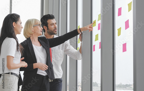 Senior glasses caucasian female manager meeting with small team and discussing on sticky notes at the window.