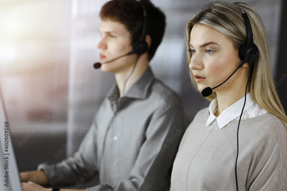 Diverse people group in sunny call center. Blonde business woman talking by headset while sitting in modern office Diverse people group in sunny call center. Blonde business woman talking by headset while sitting in modern office