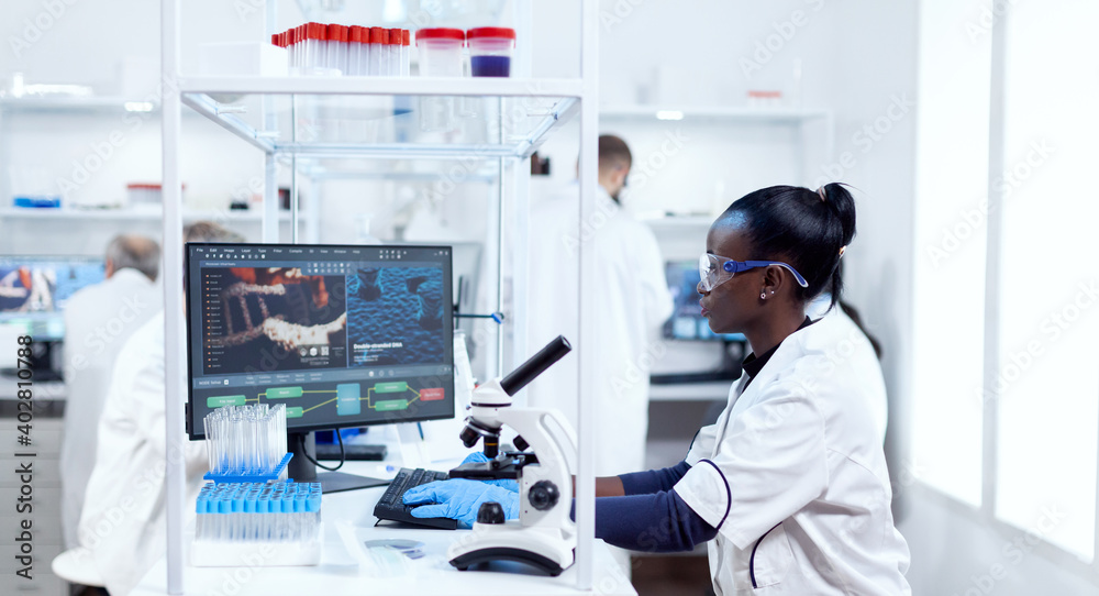 African scientist working on computer in modern facility and colleagues in the background. Black healthcare reseacher in biochemistry laboratory wearing sterile equipment.