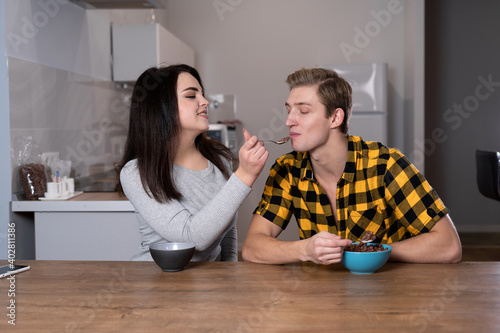 young Caucasian couple having breakfast at home. eat cereals with milk sitting at the table in the kitchen. They feed each other.