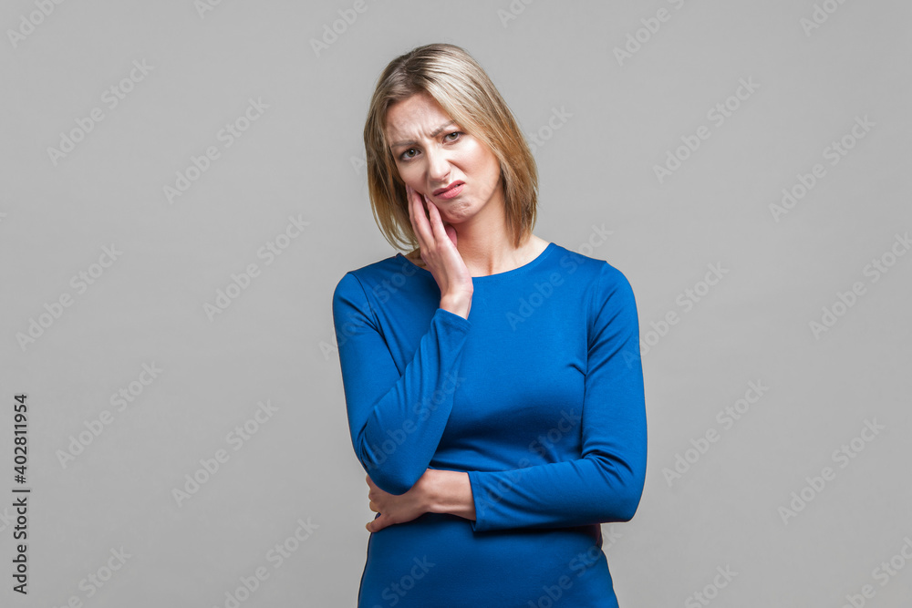 I'm bored! Portrait of indifferent thoughtful woman in elegant tight blue dress keeping hand on cheek and looking disinterested and dissatisfied. indoor studio shot isolated on gray background I'm bored! Portrait of indifferent thoughtful woman in elegant tight blue dress keeping hand on cheek and looking disinterested and dissatisfied. indoor studio shot isolated on gray background