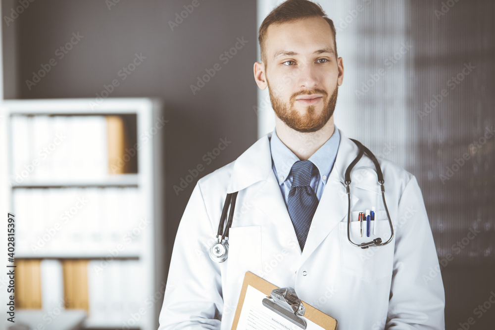 Red-bearded doctor standing straight in clinic near his working place. Portrait of physician. Medicine and healthcare concept