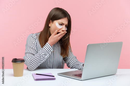 Tired sleepy woman yawning closing mouth with hand, working on laptop sitting at home office and wearing eyes patches. Indoor studio shot isolated on pink background photo