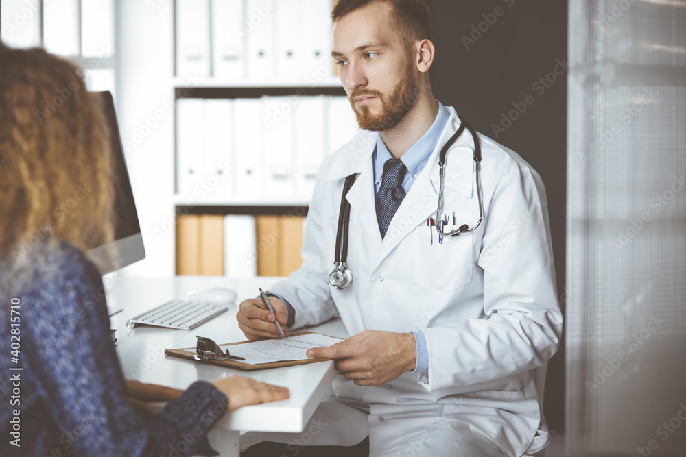 Friendly red-bearded doctor and patient woman discussing current health examination while sitting in sunny clinic. Medicine concept