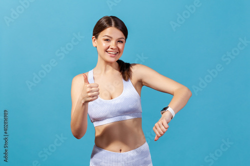 Happy athletic woman in white sportswear showing thumbs up demonstrating smartwatch on her wrist, checking indicators, satisfied with workout results. Indoor studio shot isolated on blue background © khosrork