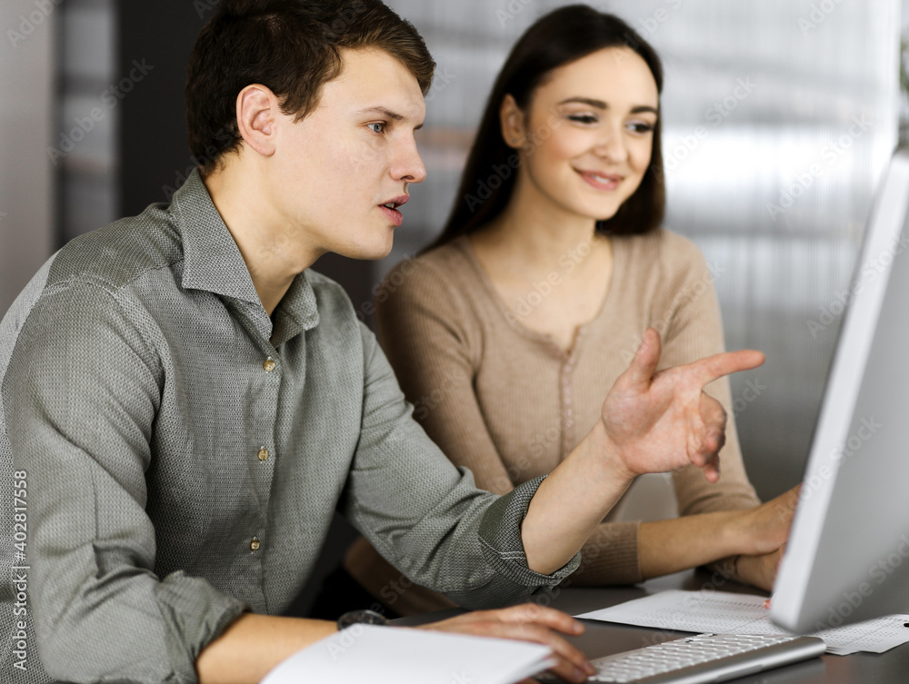 Friendly young businessman and programmer in a green shirt is working on computer, while sitting together at the desk with a female colleague in a modern office. Focus on man. Concept of successful Friendly young businessman and programmer in a green shirt is working on computer, while sitting together at the desk with a female colleague in a modern office. Focus on man. Concept of successful