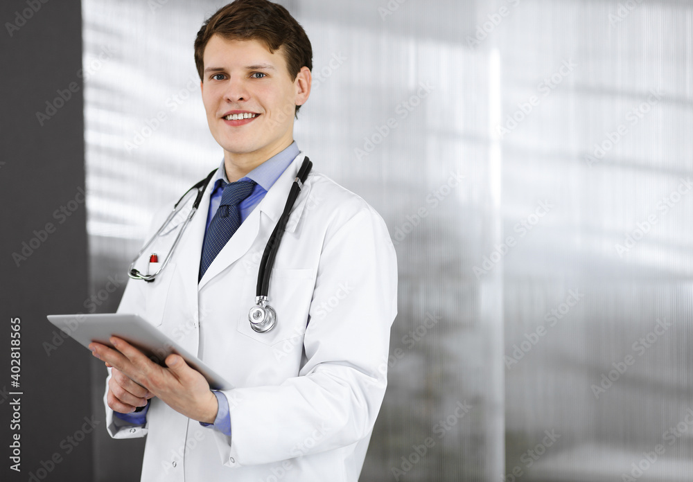 Young friendly doctor is checking some information on his computer tablet. Portrait of a professional physician at work in a clinic. Medicine concept