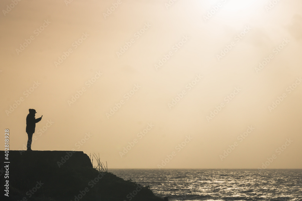 silhouette en haut d'une falaise en train de regarder son portable face à la mer