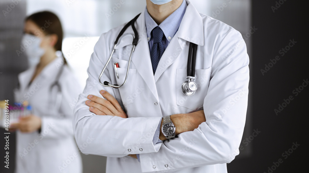 Unknown male doctor in protective mask standing with arms crossed in clinic. Medicine concept during Coronavirus pandemic. Covid 2019 Unknown male doctor in protective mask standing with arms crossed in clinic. Medicine concept during Coronavirus pandemic. Covid 2019