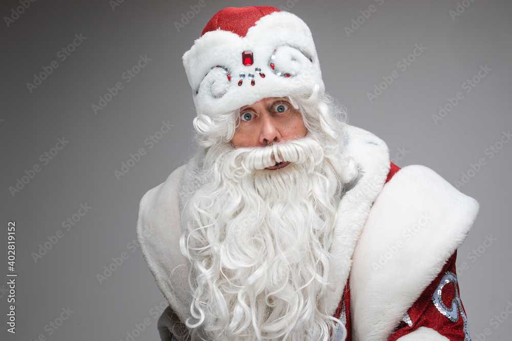 Stock photo of astonished Santa Claus in fancy costume with white long beard looking at camera with wide eyes. Isolate on grey.