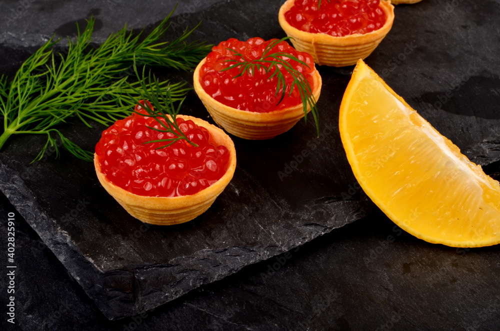 Tartlets with red salmon caviar on black stone plate, decorated with dill and a slice of lemon, close - up. Seafood appetizer Tartlets with red salmon caviar on black stone plate, decorated with dill and a slice of lemon, close - up. Seafood appetizer