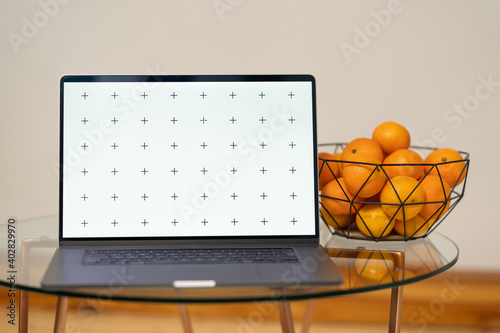 Laptop's front view mock-up with tangerines in a triangluar black hipster bowl on a glass table, with a plant in background, blank screen for your design, with free space for your text about business photo