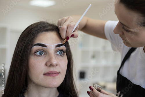 The master draws auxiliary lines between the eyebrows with a white pencil for permanent makeup. Creating an eyebrow sketch. Correction and coloring of eyebrows.