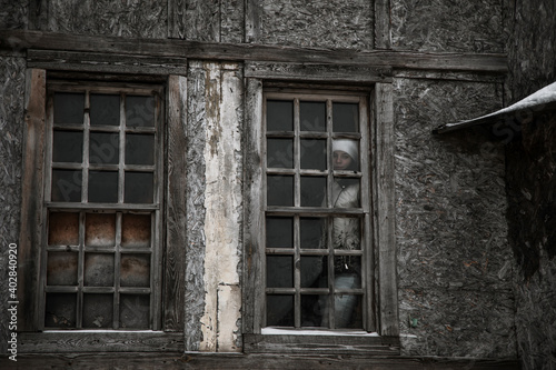 The girl stands outside the window in an old abandoned house. Girl in a white jacket. Dilapidated and old window. © Kooper