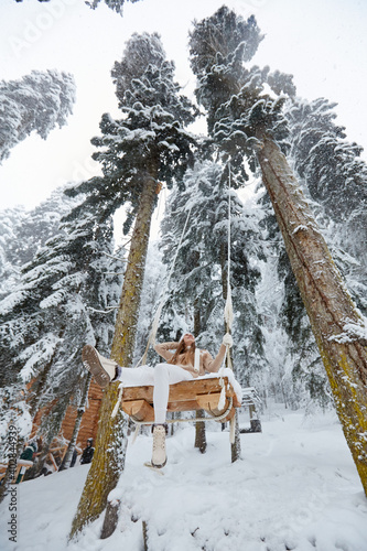 Young girl swinging on a long swing on snow forest mountain view. Swinging between tall pines. Bottom-up view