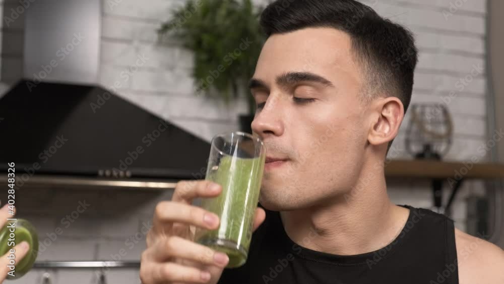 Making green juice at home: Young sporty man in sportswear drinking smoothie from a glass. Close-up shot. Healthy breakfast after a workout in the kitchen 4k