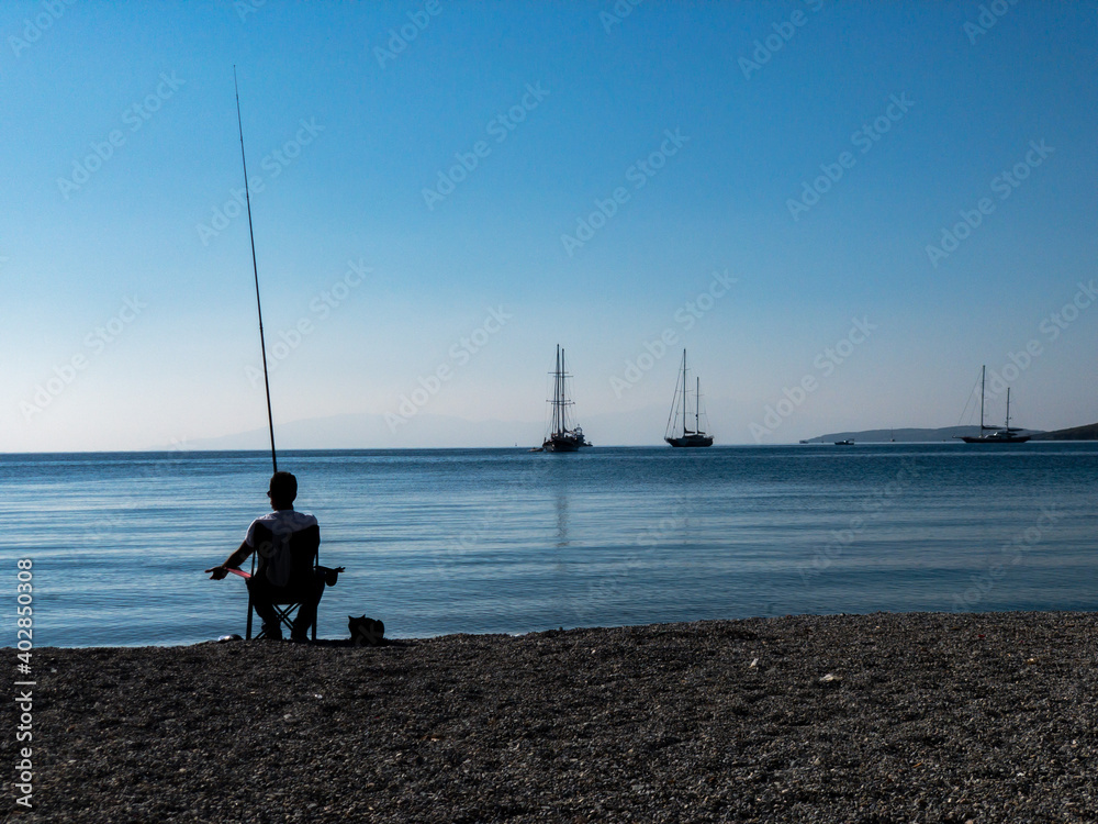 A lonely fisherman silhouette sits in a chair at the beach and tries to catch some fishes. A cat lies beside him and waiting for food. Some sailing boats are on anchor. Blue sky and sea background
