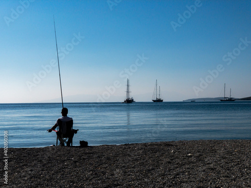 A lonely fisherman silhouette sits in a chair at the beach and tries to catch some fishes. A cat lies beside him and waiting for food. Some sailing boats are on anchor. Blue sky and sea background