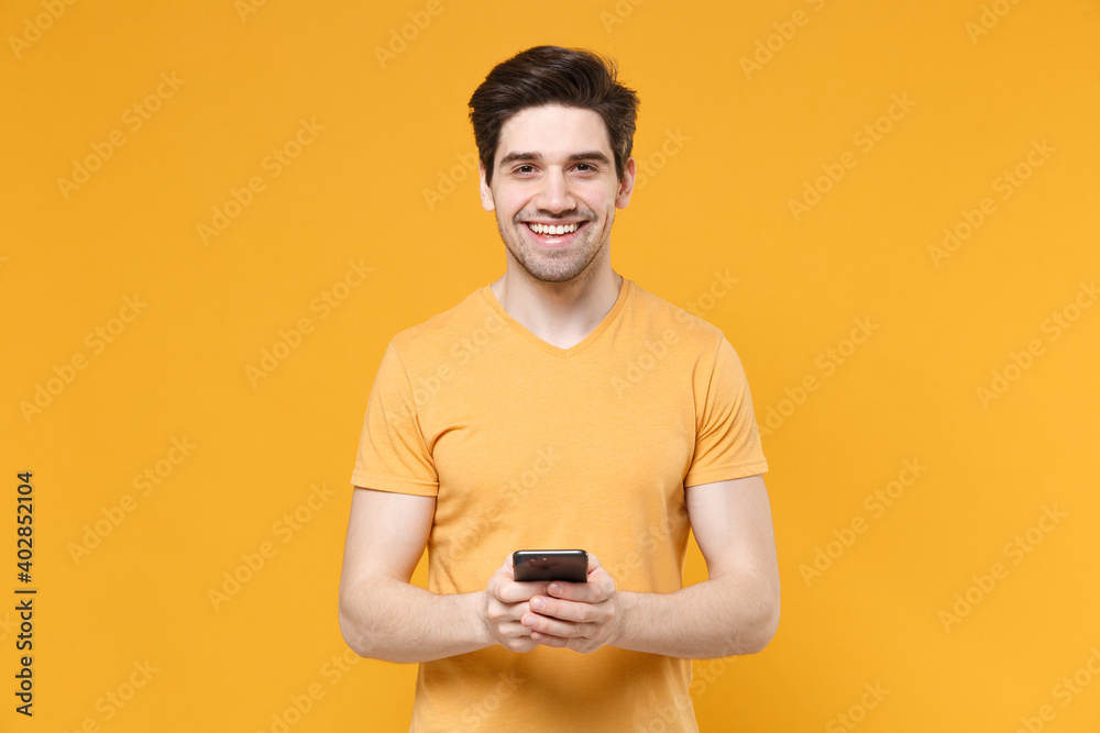 Young smiling unshaved caucasian student man 20s wearing casual basic blank print design t-shirt holding modern mobile cell phone looking camera isolated on yellow color background studio portrait