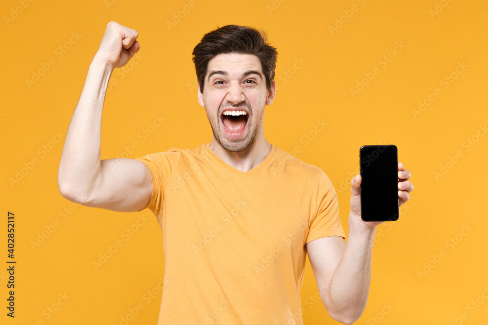Young satisfied man in casual print t-shirt hold mobile cell phone with blank screen workspace copy space mock up doing winner gesture clenching fists isolated on yellow background studio portrait. Young satisfied man in casual print t-shirt hold mobile cell phone with blank screen workspace copy space mock up doing winner gesture clenching fists isolated on yellow background studio portrait.