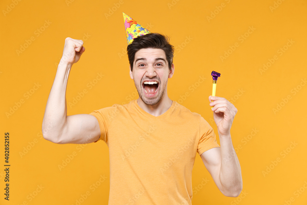 Young unshaved cute happy excited man 20s in basic t-shirt wearing birthday hat cone celebrating holding pipe doing winner gesture with clenching fist isolated on yellow background studio portrait. Young unshaved cute happy excited man 20s in basic t-shirt wearing birthday hat cone celebrating holding pipe doing winner gesture with clenching fist isolated on yellow background studio portrait.