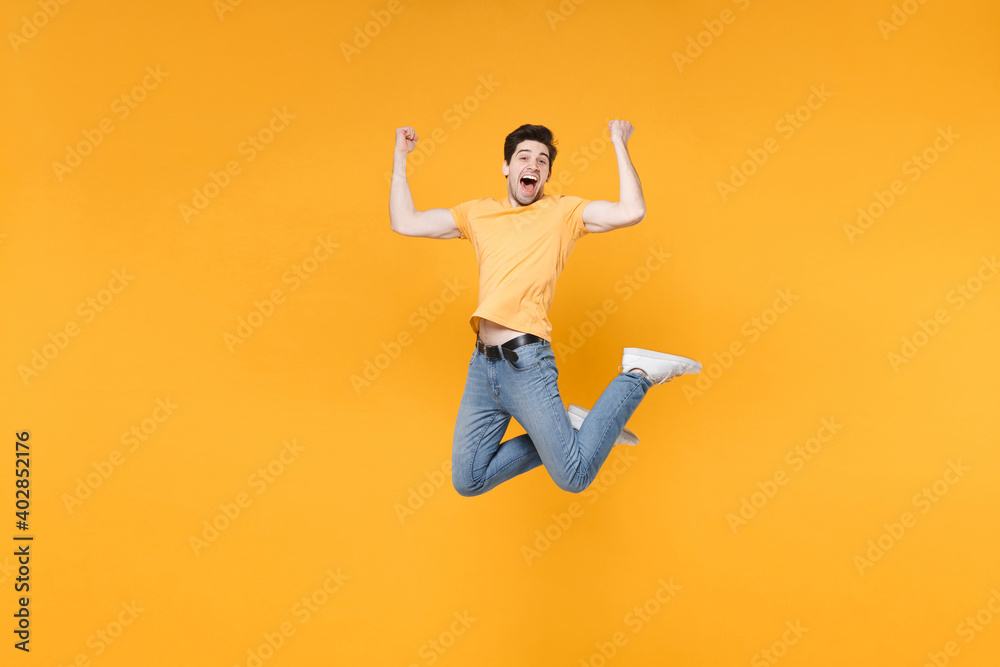 Full length of young happy successful overjoyed excited fun surprised man in casual t-shirt jeans high jump up doing winner gesture with clenching fists isolated on yellow background studio portrait.