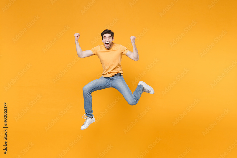 Fototapeta premium Full length young successful caucasian fun happy surprised man 20s wearing casual t-shirt jeans high jumping up doing winner gesture clenching fists isolated on yellow background studio portrait