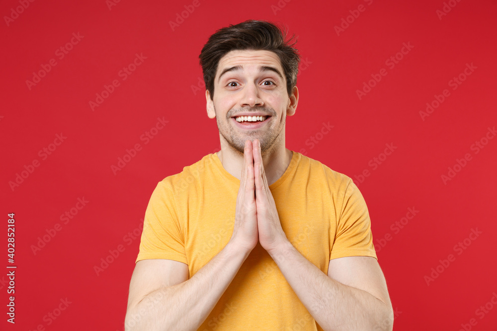 Young unshaved wishful tustful expecting caucasian smioling man 20s wearing casual basic yellow t-shirt look camera holding hands in prayer gesture isolated on red color background studio portrait.