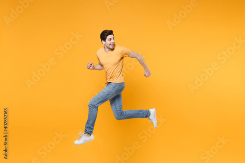Full length side view of young caucasian fun happy energetic hurrying man 20s wearing casual basic t-shirt jeans high jumping up running look back isolated on yellow color background studio portrait.