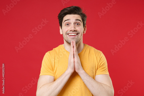 Young unshaved wishful tustful expecting caucasian smioling man 20s wearing casual basic yellow t-shirt look camera holding hands in prayer gesture isolated on red color background studio portrait. photo