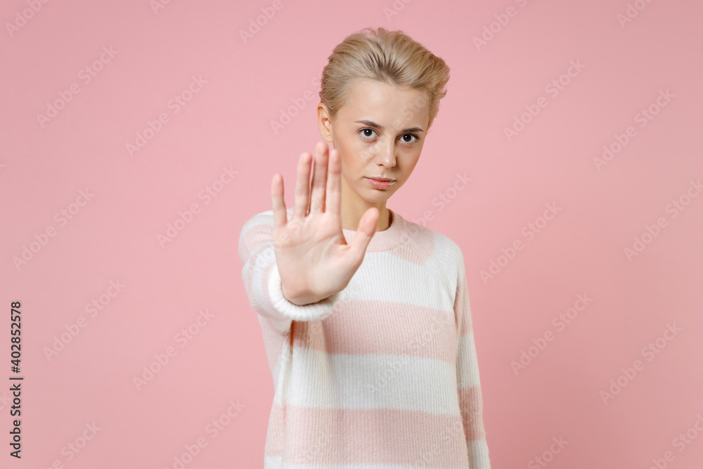Fototapeta premium Young serious blonde woman 20s with short haircut nude makeup in casual knitted sweater show stop gesture with palm keep social distance look camera isolated on pastel pink background studio portrait.