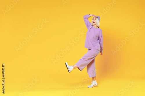 Full length of young fun woman 20s bob haircut in casual basic purple suit beanie hat walk raised leg hold hand at forehead look far away distance isolated on yellow color background studio portrait.