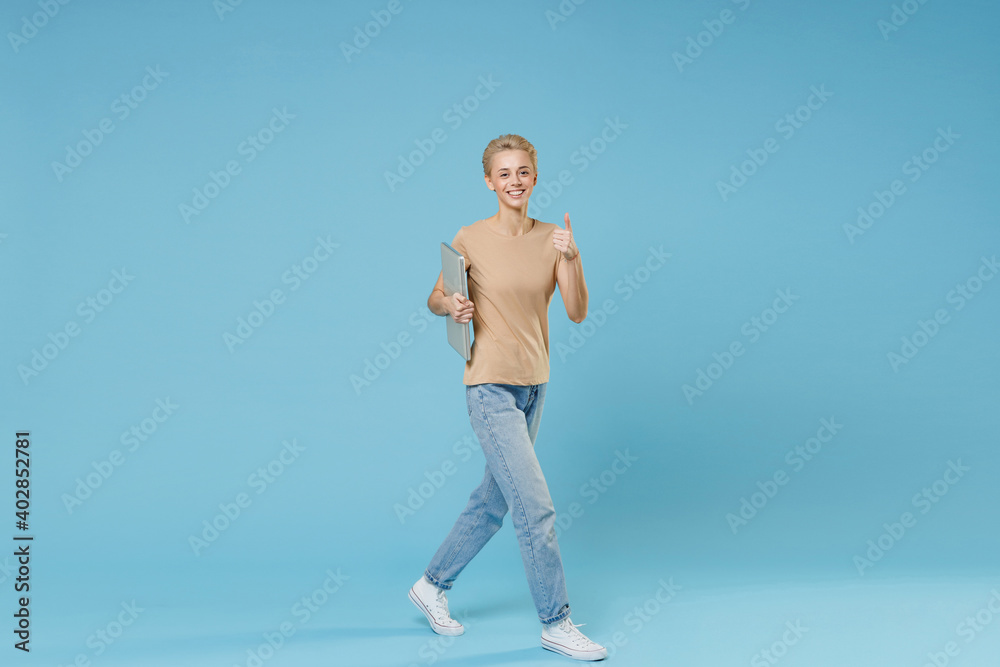Full length of smiling caucasian young woman 20s short haircut wear casual beige t-shirt jeans walk with closed laptop pc computer show thumb up gesture isolated on blue background studio portrait