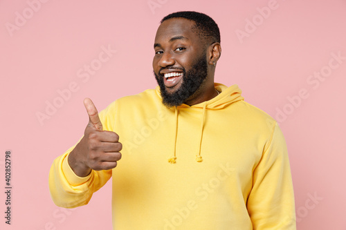 Smiling cheerful funny young african american man 20s wearing casual basic yellow streetwear hoodie standing showing thumb up looking camera isolated on pastel pink color background studio portrait.